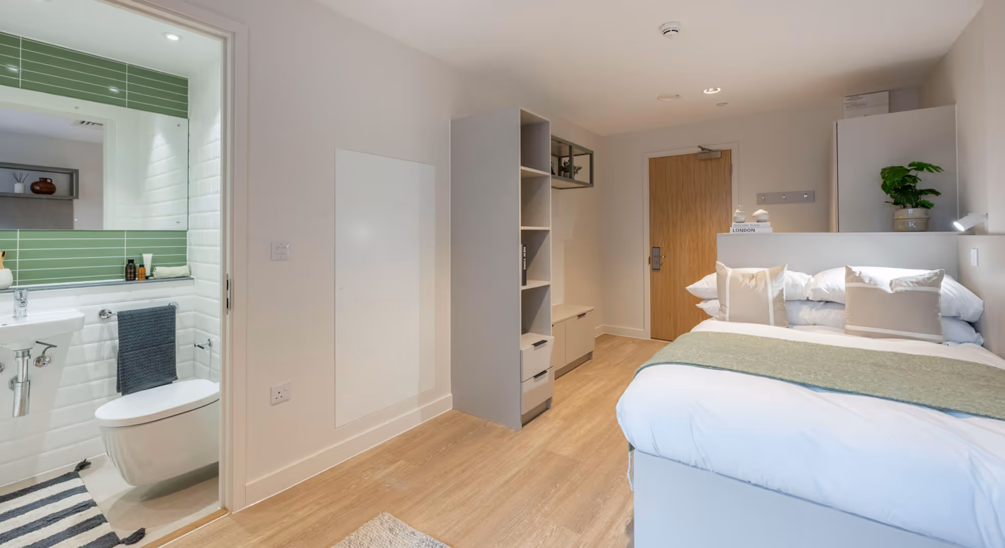 Modern bedroom with wooden floor, a bed with white and beige pillows, a green throw, shelving unit, and an ensuite bathroom with green and white tiled walls.