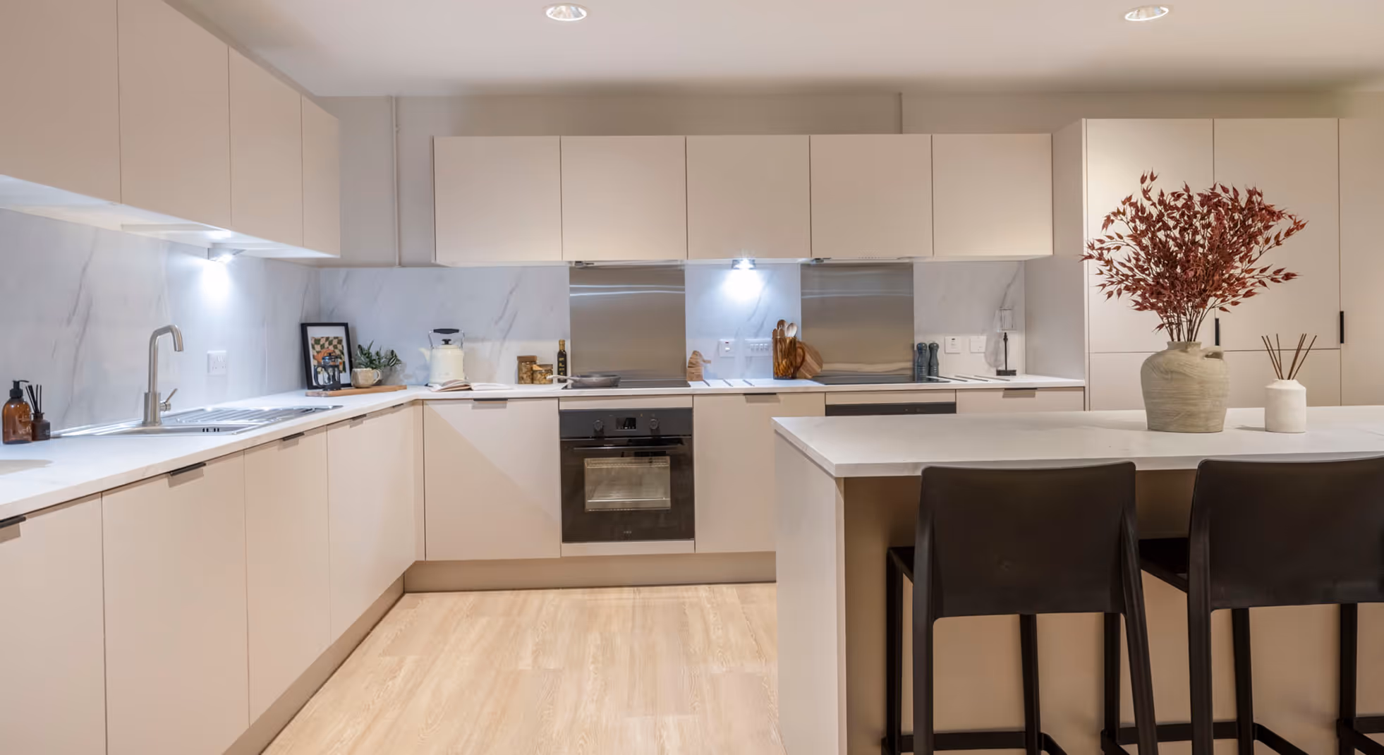 Modern kitchen with white cabinets, marble countertop, stainless steel oven and backsplash, kitchen island with black stools, and decorative vases with plants.