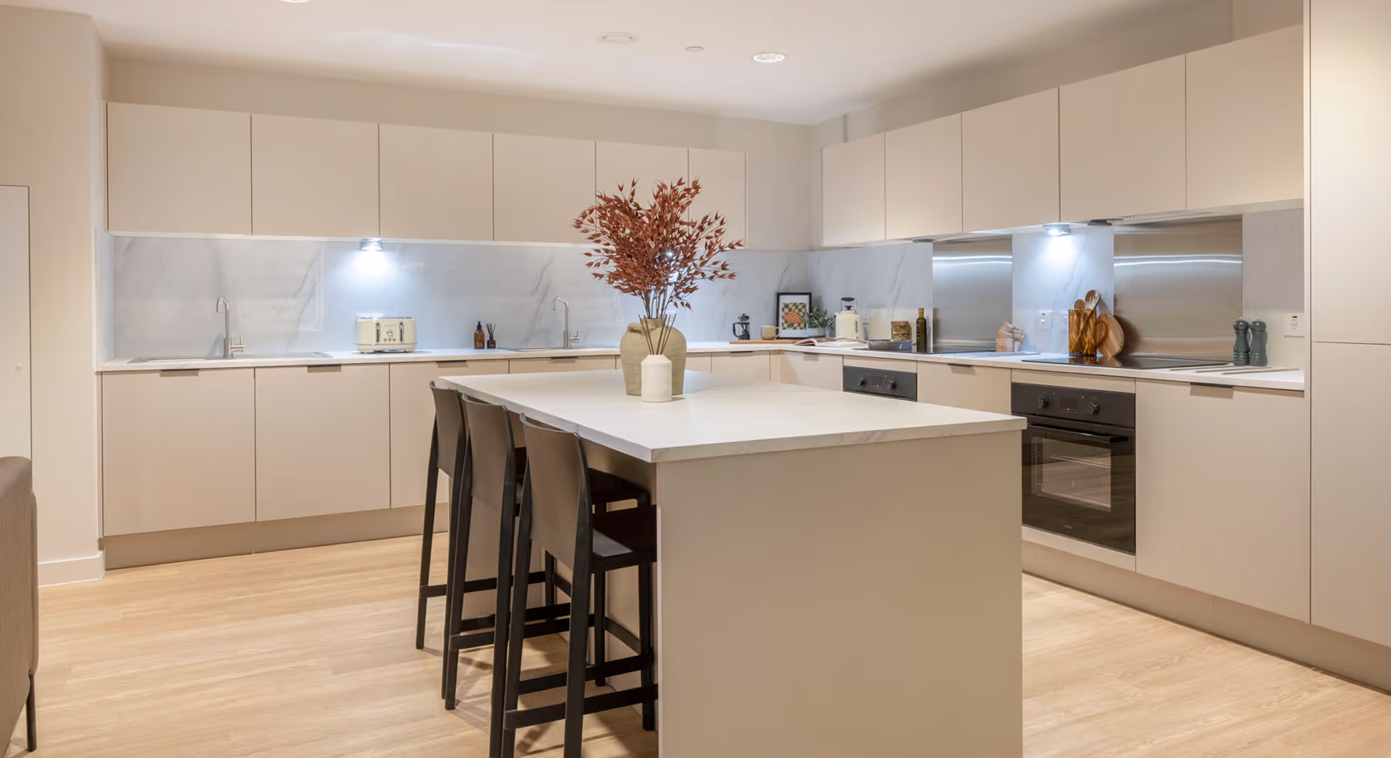 Modern kitchen with beige cabinets, marble backsplash, central island with black bar stools, and decorative vases with dried branches.