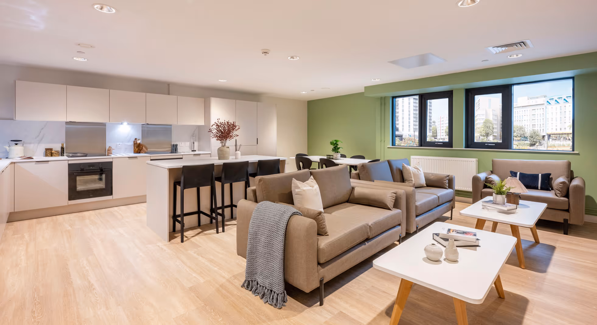 Modern open-plan living room and kitchen with beige sofas, white tables, and large windows overlooking city buildings.