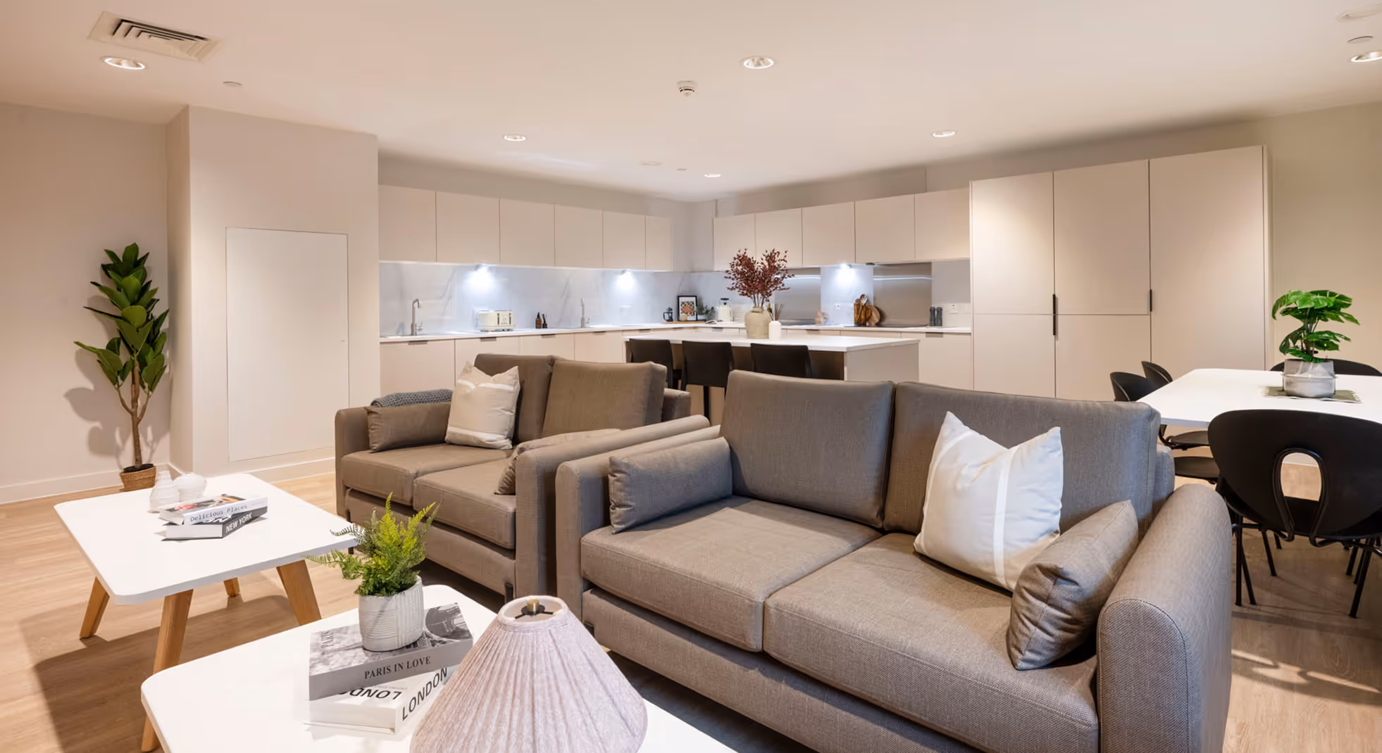 Modern open-concept living area with beige sofas, white coffee tables, and a well-lit kitchen with cream cabinetry and a dining table with black chairs.