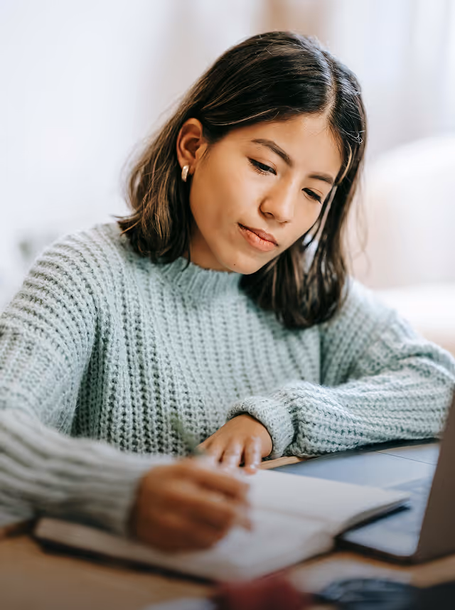 Young woman in a light blue sweater writing in a notebook with a laptop nearby.