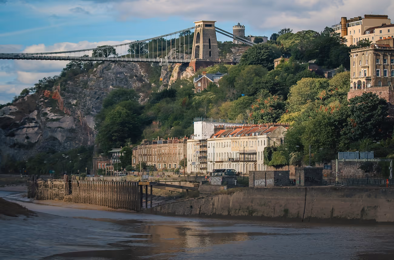 Clifton Suspension Bridge spanning rocky cliffs with residential buildings and trees along the riverbank under a partly cloudy sky.