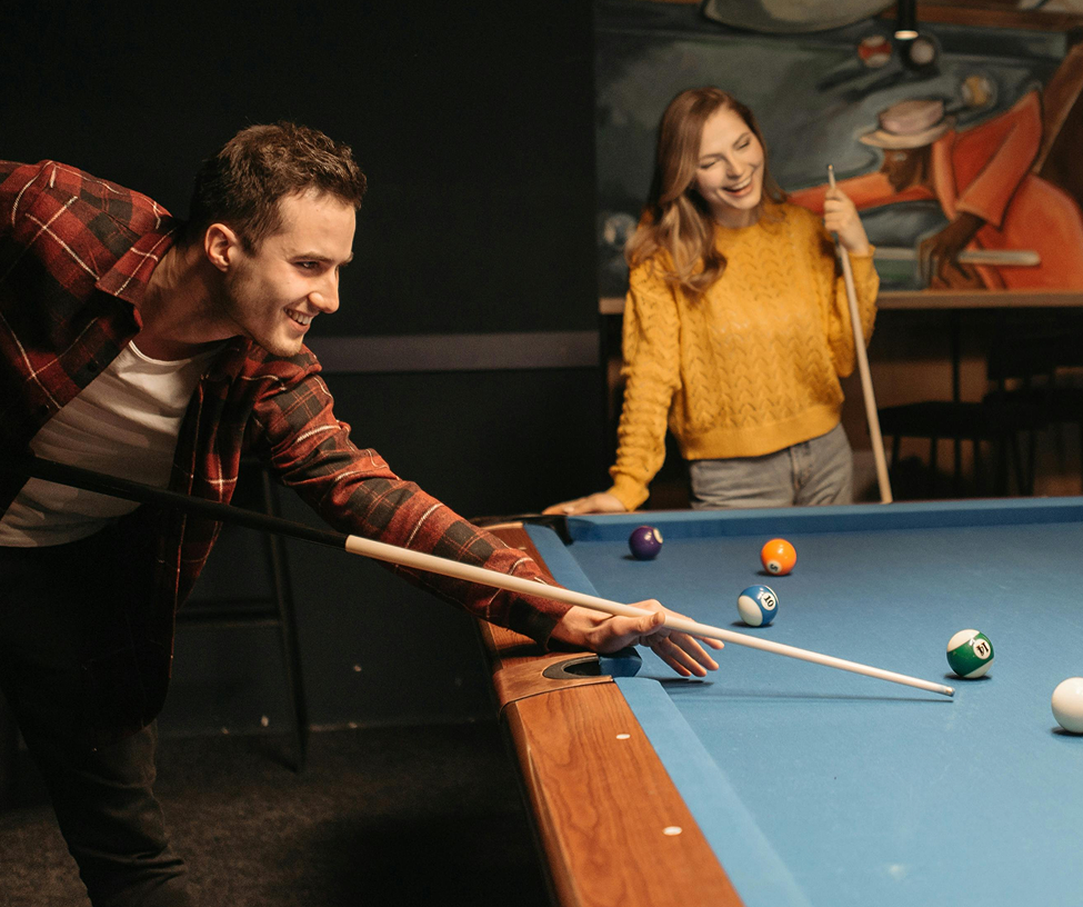 Man in a red plaid shirt aiming a pool cue on a billiards table while a woman in a yellow sweater watches and smiles.