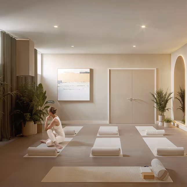 Quiet yoga studio with six mats and bolsters arranged evenly, a woman in white sitting thoughtfully on one mat, soft natural light, neutral tones, plants, and abstract wall art.