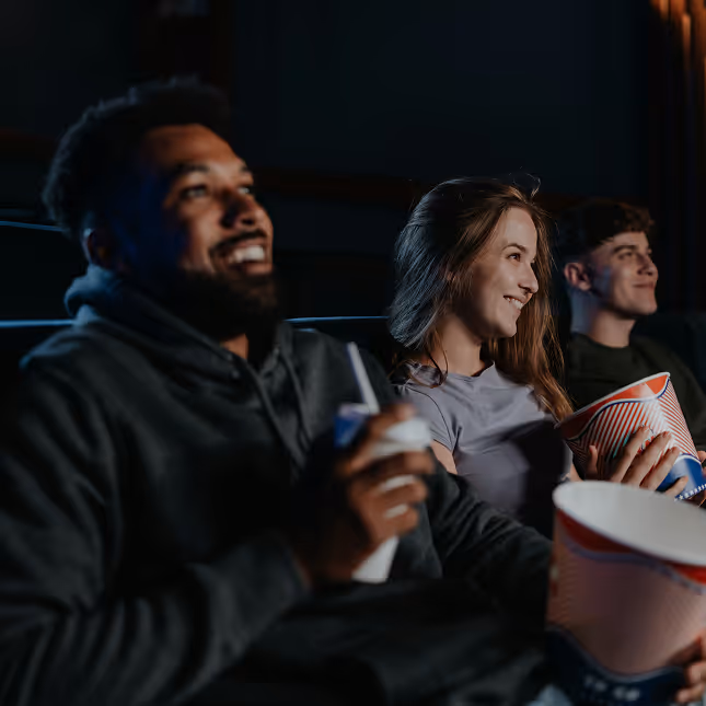 Smiling young people sitting in a dark movie theater holding popcorn and drinks.