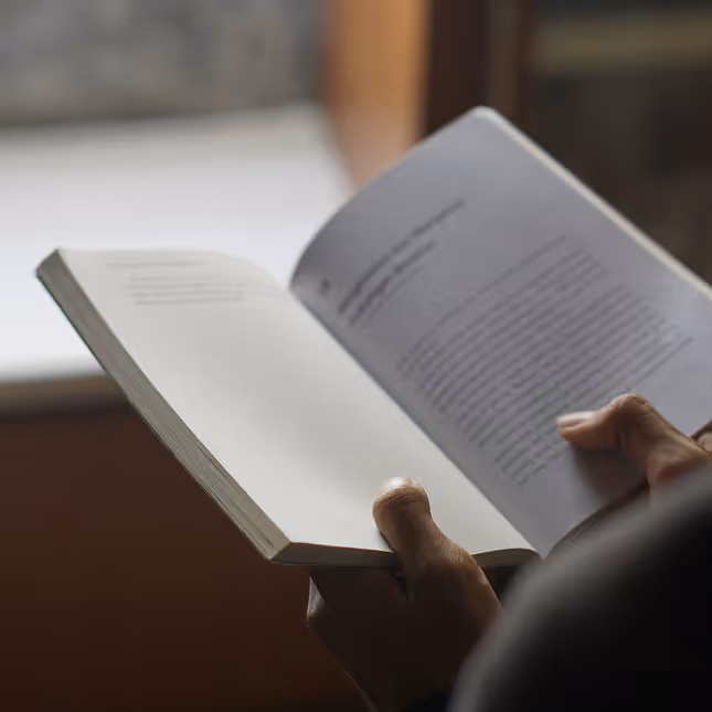 Close-up of a person holding and reading an open book indoors near a window.
