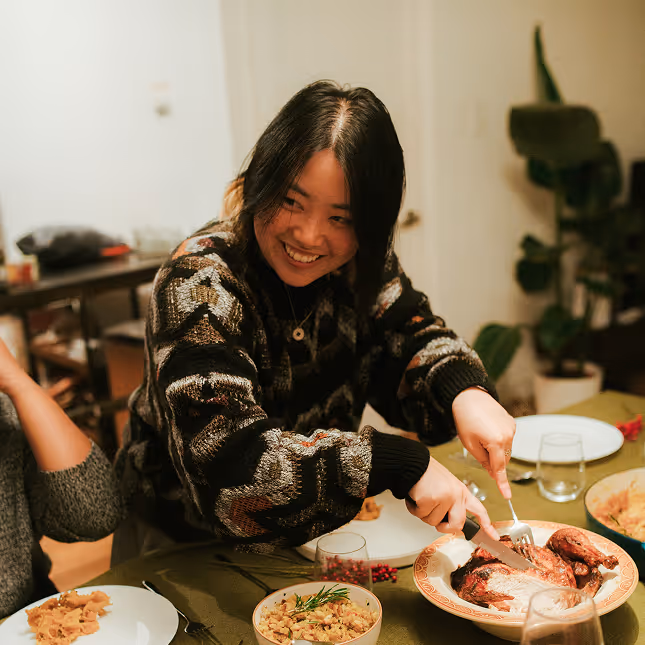 Smiling woman with dark hair carving a roasted chicken at a dinner table set with dishes and glasses.