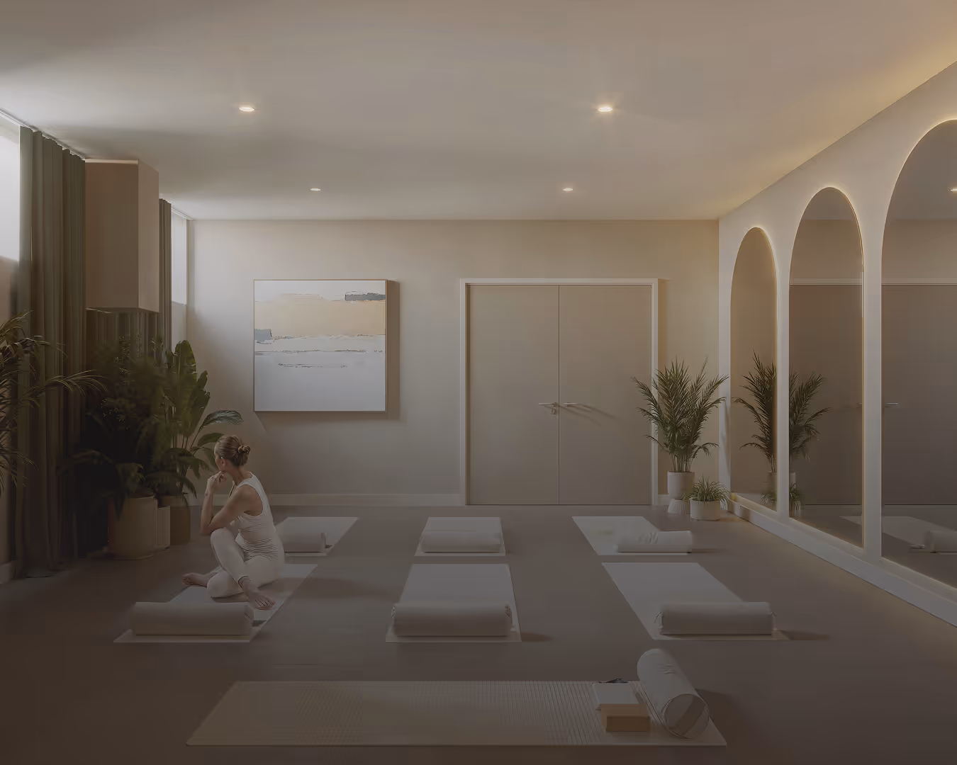 Calm yoga studio with six mats neatly arranged on the floor, a woman sitting cross-legged on one mat, neutral tones, large wall mirrors, plants, and soft lighting.