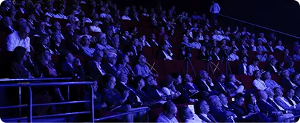 Un public nombreux assis dans une salle sombre, regardant une présentation ou conférence.