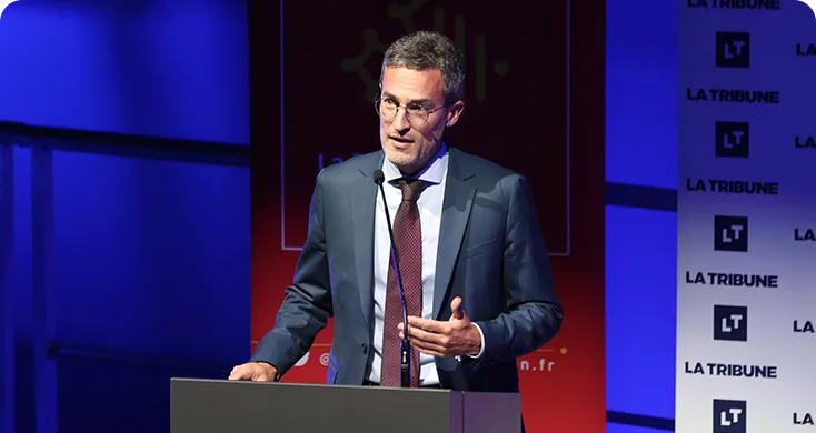 Un homme en costume parle au micro lors d'une conférence avec un fond bleu et rouge.