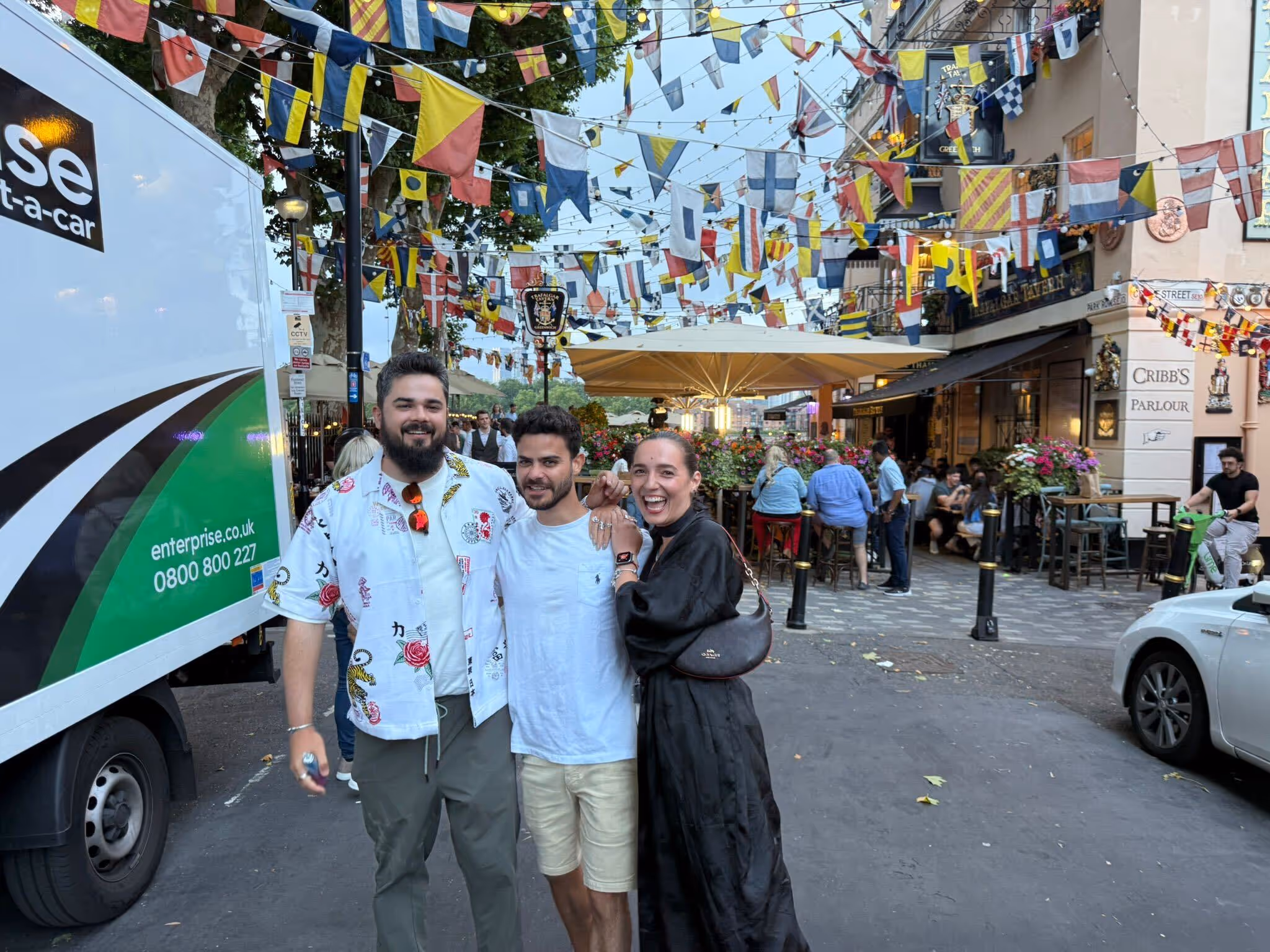 Three friends smiling and posing together on a street decorated with colorful international flags and outdoor seating at Cribb's Parlour.