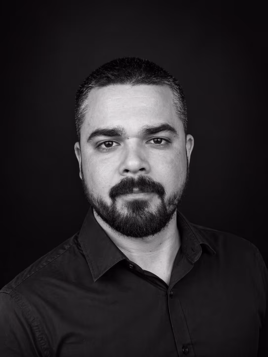 Black and white portrait of a man with short hair and a beard wearing a dark collared shirt against a dark background.