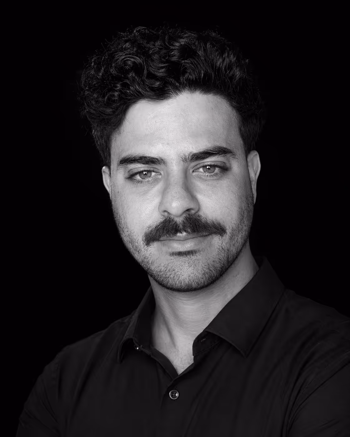 Black and white portrait of a man with curly hair, a mustache, and light stubble, wearing a dark button-up shirt against a black background.