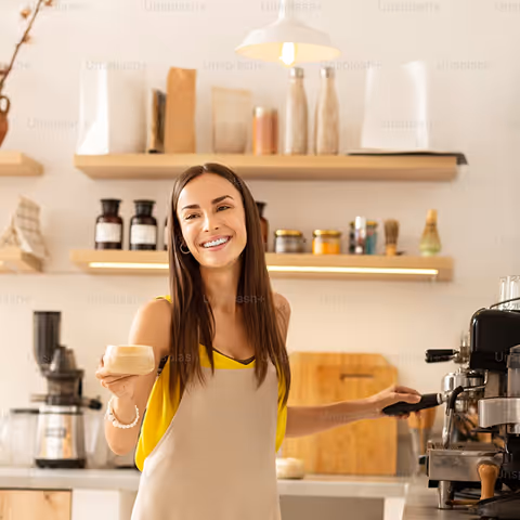 Smiling woman in a kitchen holding a coffee cup and operating an espresso machine.