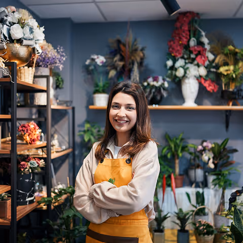 Smiling woman in a yellow apron standing with arms crossed inside a flower shop surrounded by plants and floral arrangements.