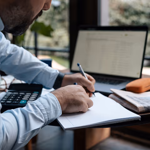 Person writing on a notepad at a desk with an open laptop and calculator nearby.
