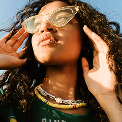 Woman with curly hair wearing large cat-eye sunglasses and layered beaded necklaces, touching her glasses while looking up.