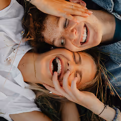 Two young women lying close together outdoors, smiling and laughing with hands gently touching their faces.