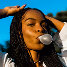 Woman with long braided hair blowing a bubble gum bubble outdoors under a clear blue sky.