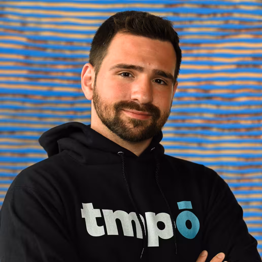Man with short dark hair and light beard wearing a light gray sweatshirt with a small astronaut emblem, standing with arms crossed against a gradient blue background.