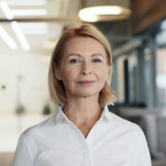 Confident middle-aged woman with short blonde hair wearing a white collared shirt in a modern office setting.