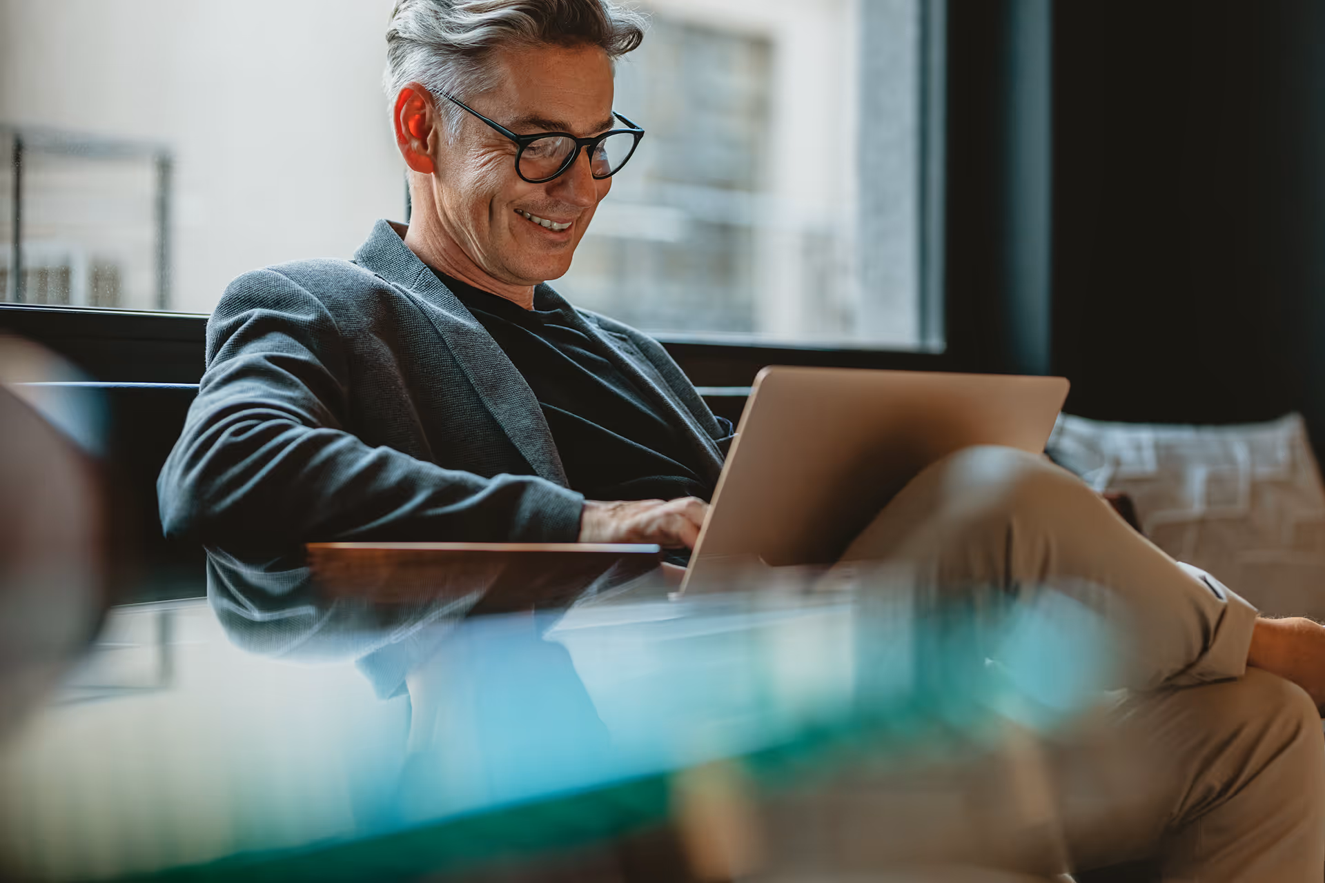 Smiling middle-aged man with gray hair and glasses using laptop while sitting comfortably indoors.