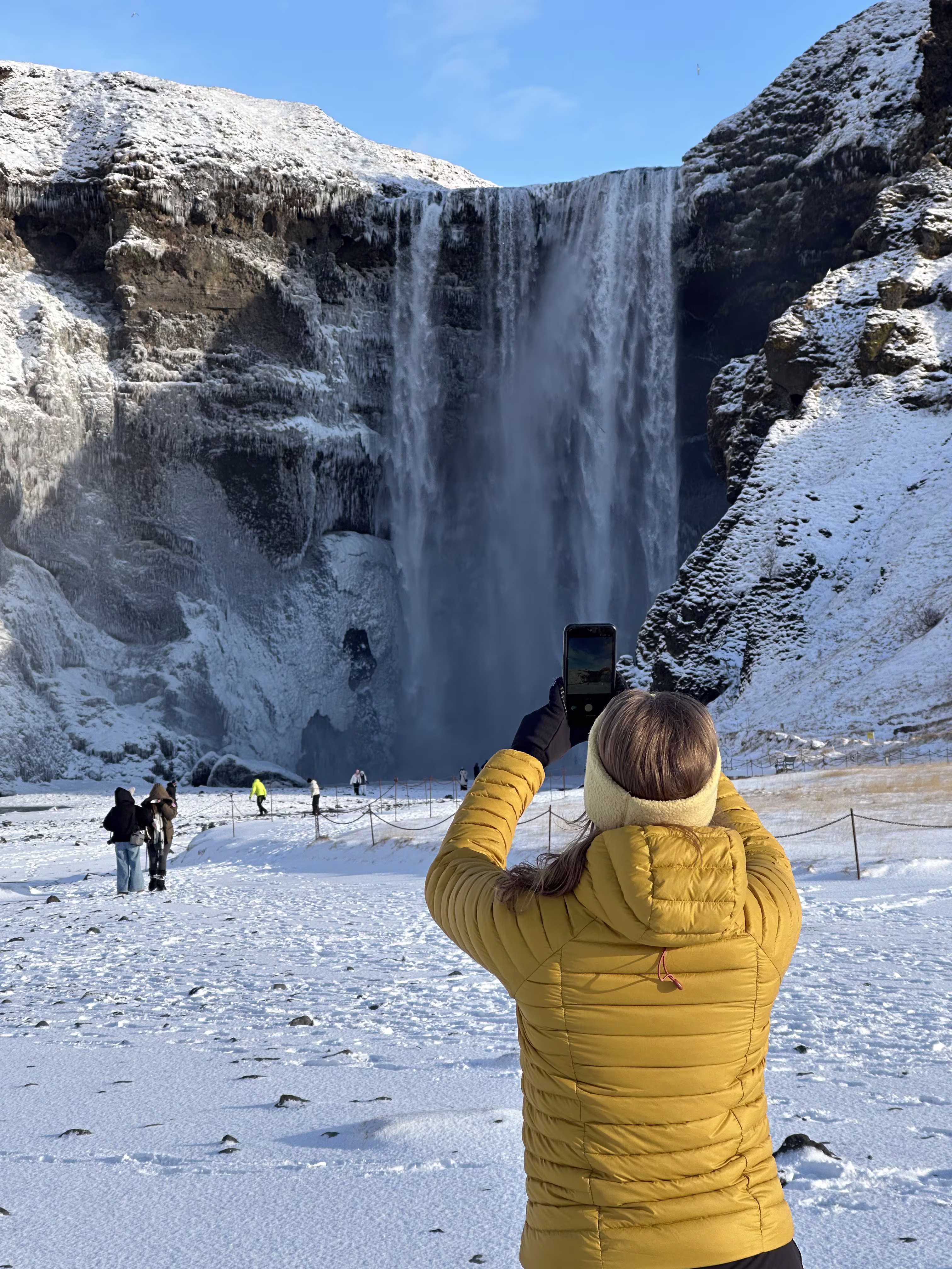 Visitor in yellow jacket photographing Skógafoss waterfall in winter