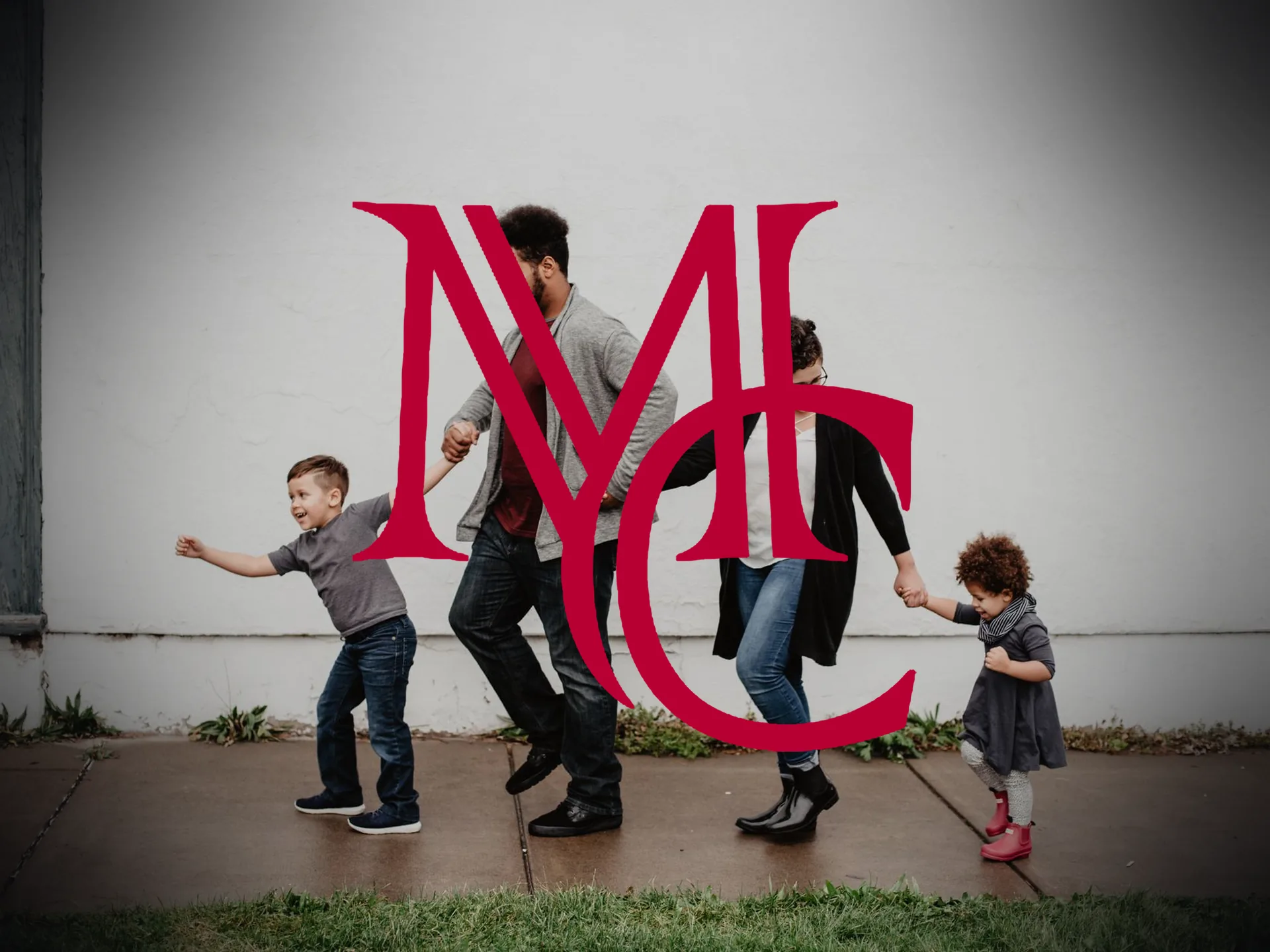 A family with two young children walking hand in hand along a sidewalk past a white wall — MCG