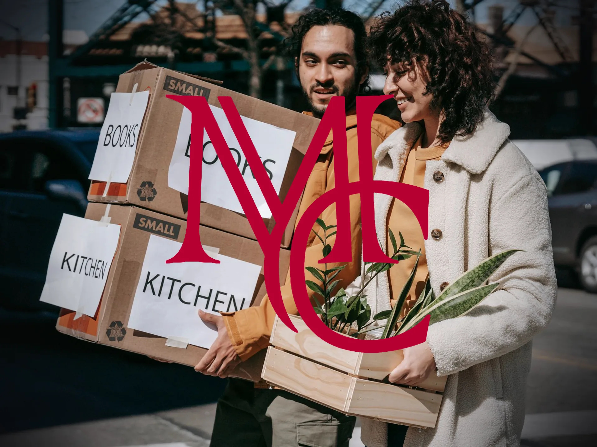 A couple carrying labeled moving boxes and a crate of houseplants along a city street — MCG