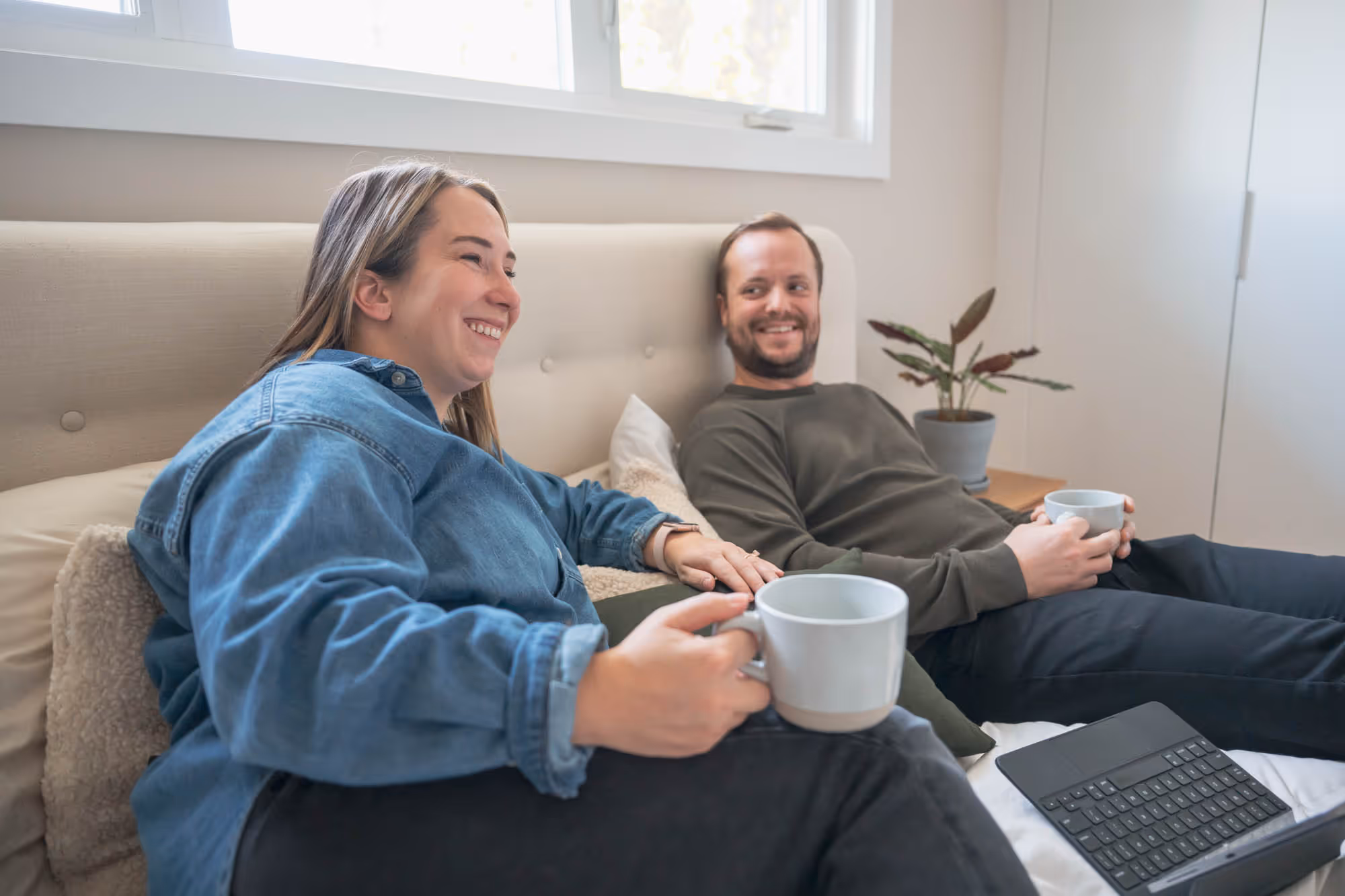 A smiling woman and man sitting comfortably on a bed with mugs, a laptop, and a potted plant nearby. Showcasing comfortable living spaces.