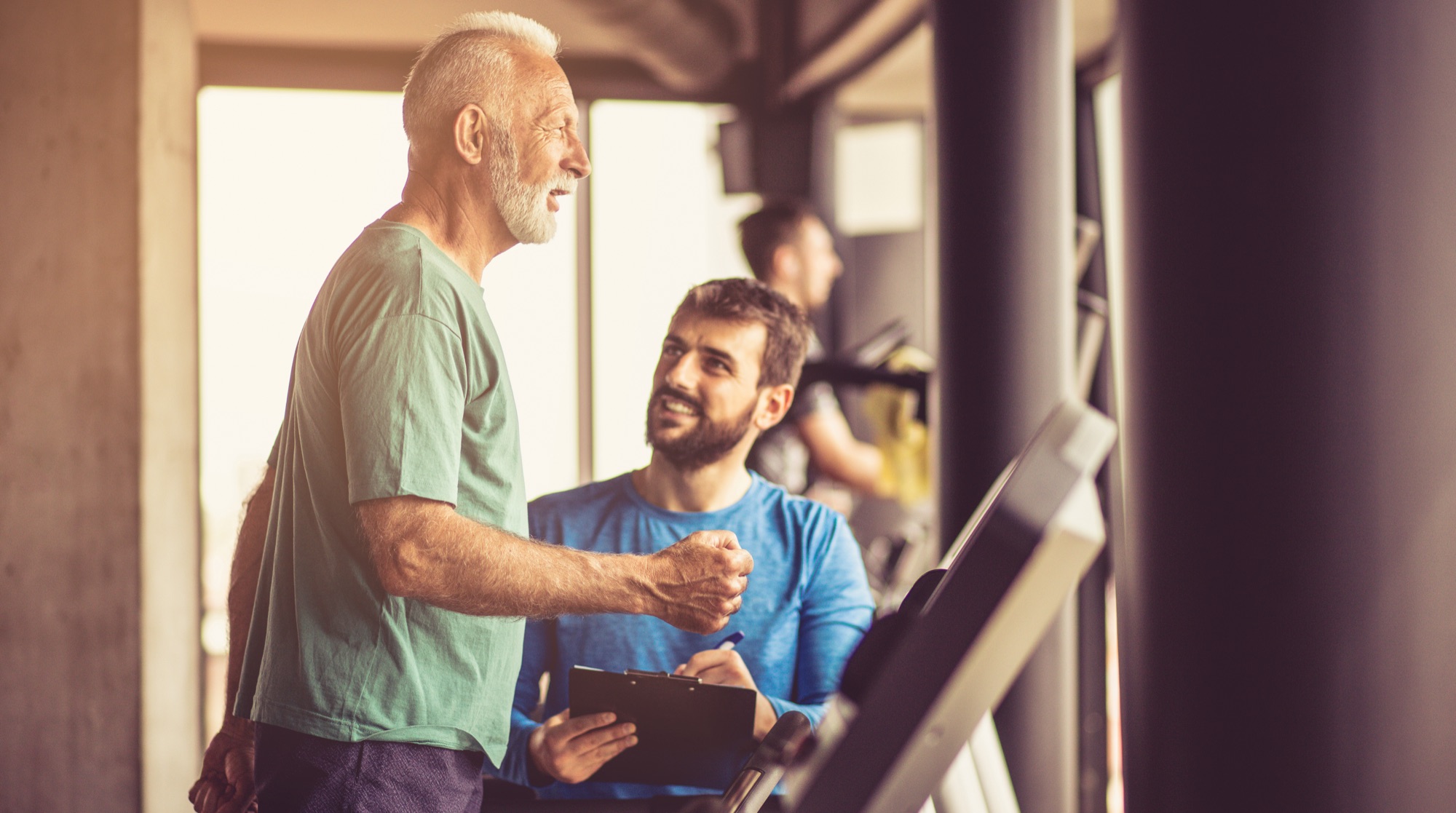 Older man talking with a rehab specialist holding a clipboard in a gym
