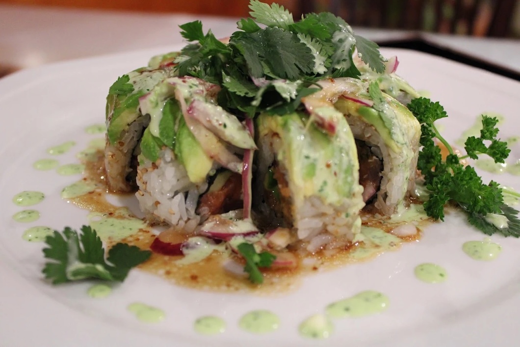 Close-up of sushi rolls topped with avocado slices, cilantro, and a creamy green sauce on a white plate.