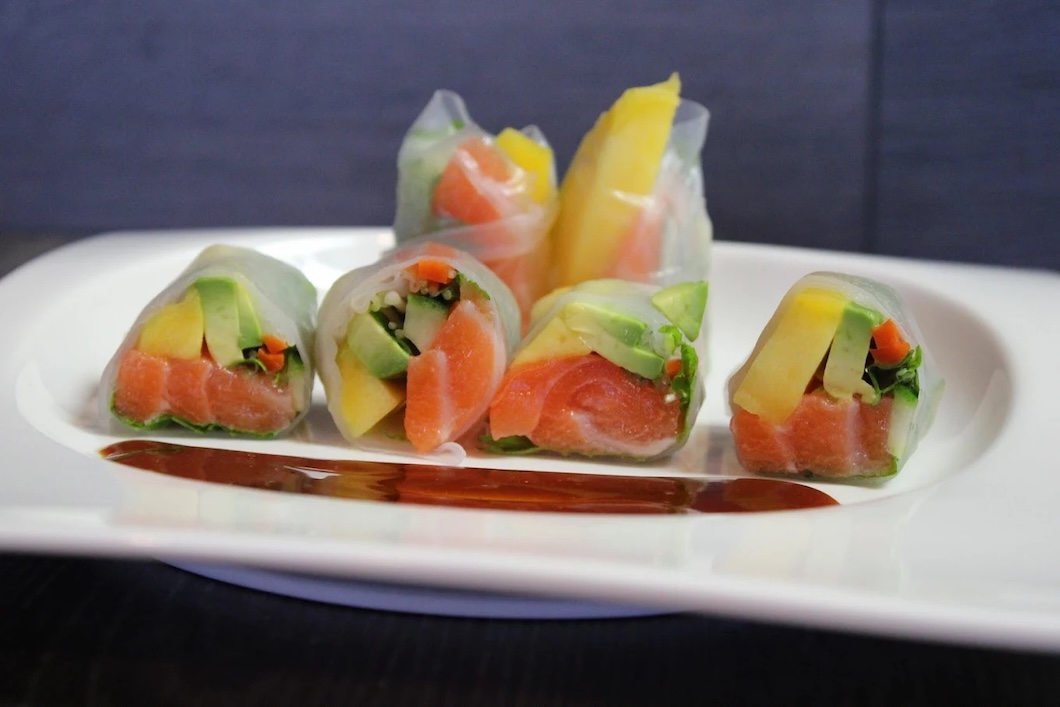 Six fresh spring rolls filled with salmon, avocado, mango, and greens on a white plate with dipping sauce.