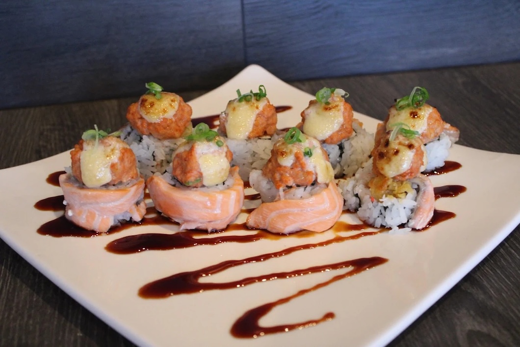 Plate of sushi rolls topped with spicy mayo, scallions, and drizzle of brown sauce on a white square dish.