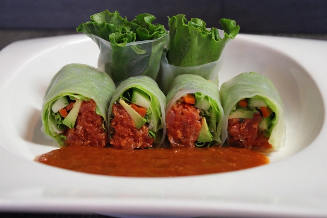 Four fresh vegetable spring rolls filled with lettuce, carrot, cucumber, and a red sauce, served with a reddish dipping sauce on a white plate.