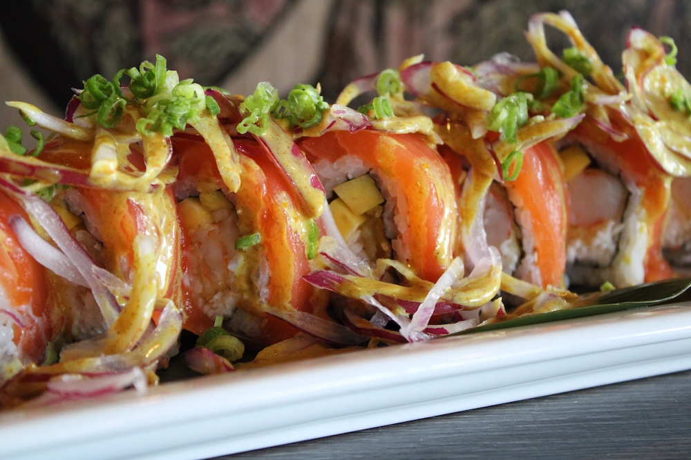 Close-up of sushi rolls topped with green onions, thinly sliced red onions, and drizzled with a creamy sauce on a white plate.