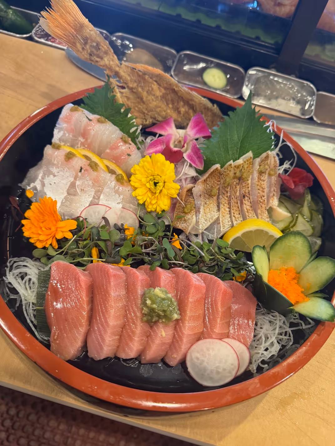 Assorted sashimi platter with various slices of raw fish, garnished with edible flowers, lemon slices, cucumber, and shredded radish.
