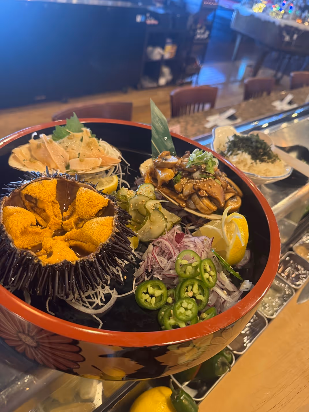 Japanese sashimi platter with sea urchin, sliced fish, mushroom-topped shell, sliced jalapeños, onions, cucumber, and lemon wedges in a decorative bowl.