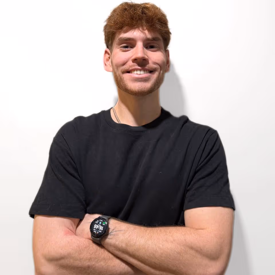 Jeune homme souriant aux cheveux roux, bras croisés, portant un t-shirt noir et une montre noire.