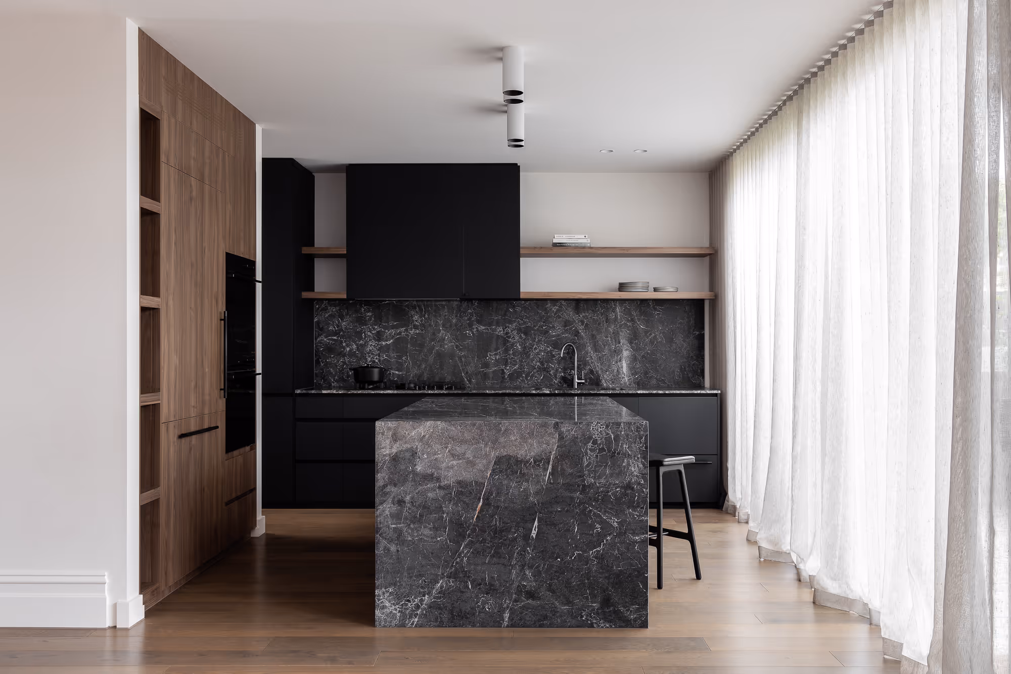 Modern kitchen with black marble island, black cabinets, wooden shelves, and large sheer white curtains over hardwood floors.