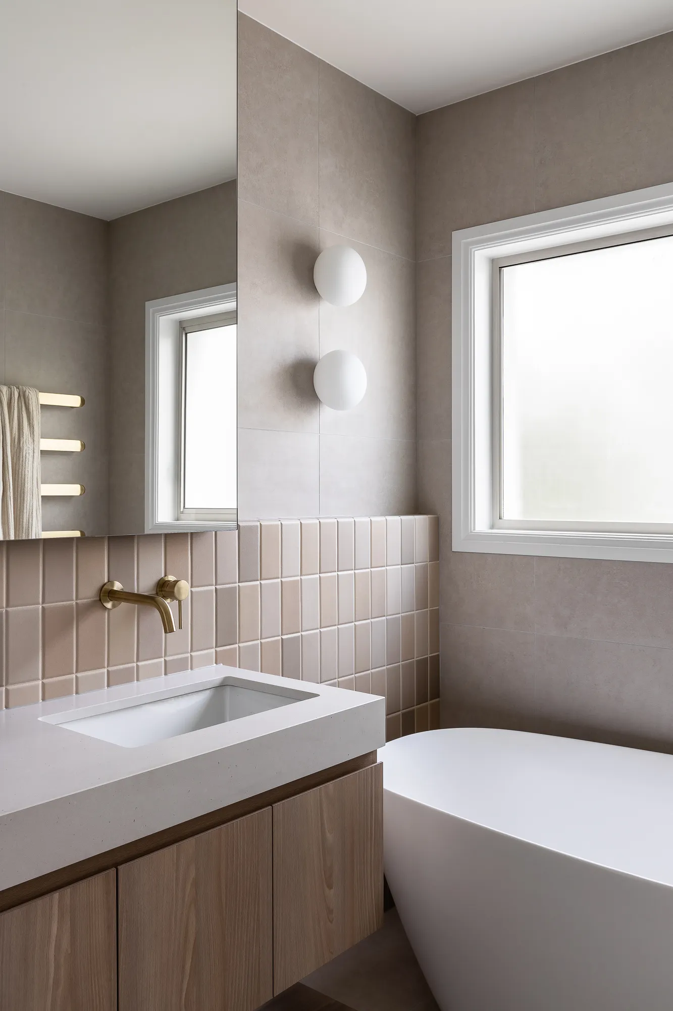 Modern bathroom with a white rectangular sink, brass wall-mounted faucet, wooden vanity, beige tiles, and a white freestanding bathtub under a frosted window.