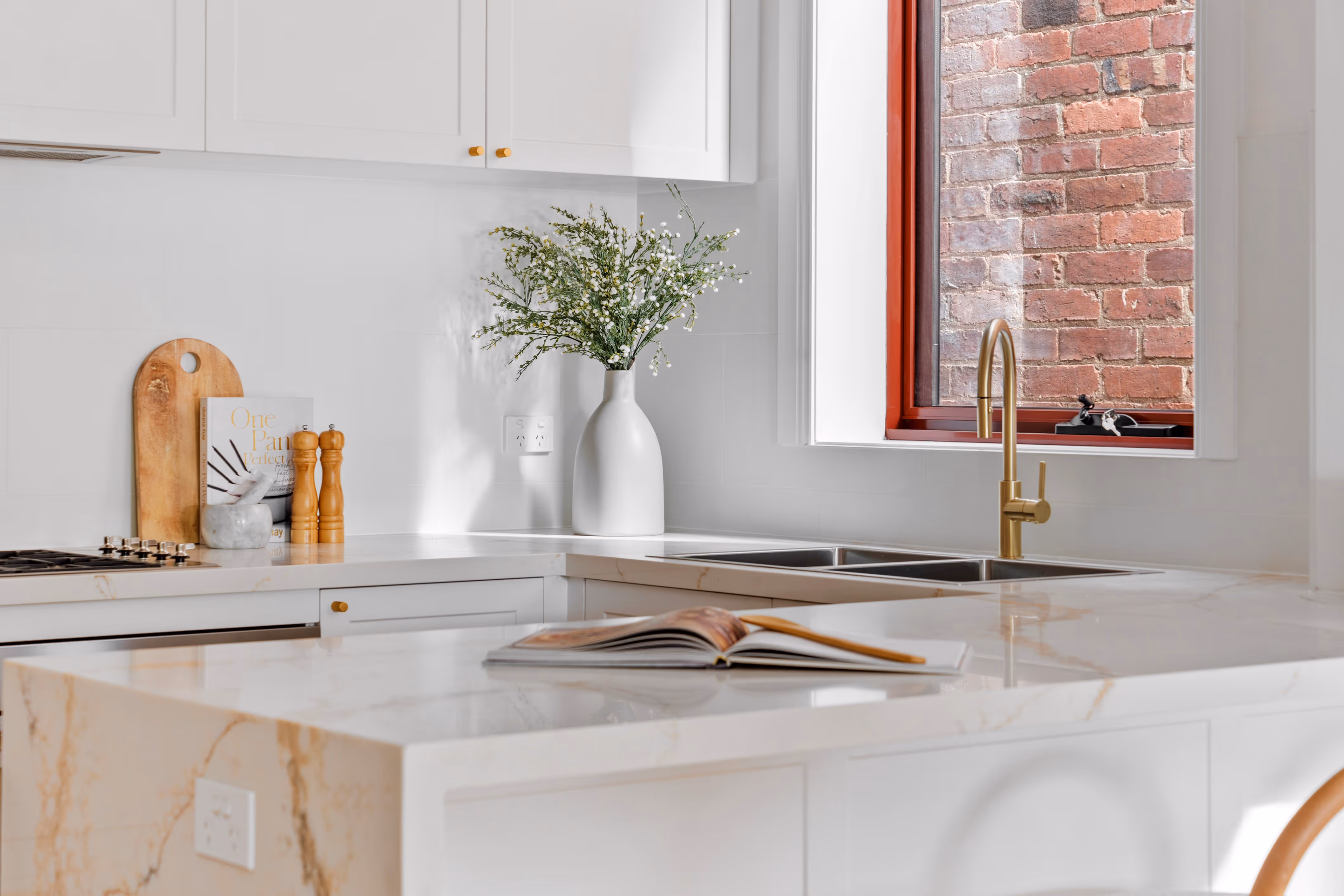 Modern white kitchen with marble countertops, gold faucet, a white vase with flowers, wooden cutting board, and an open cookbook on the island.