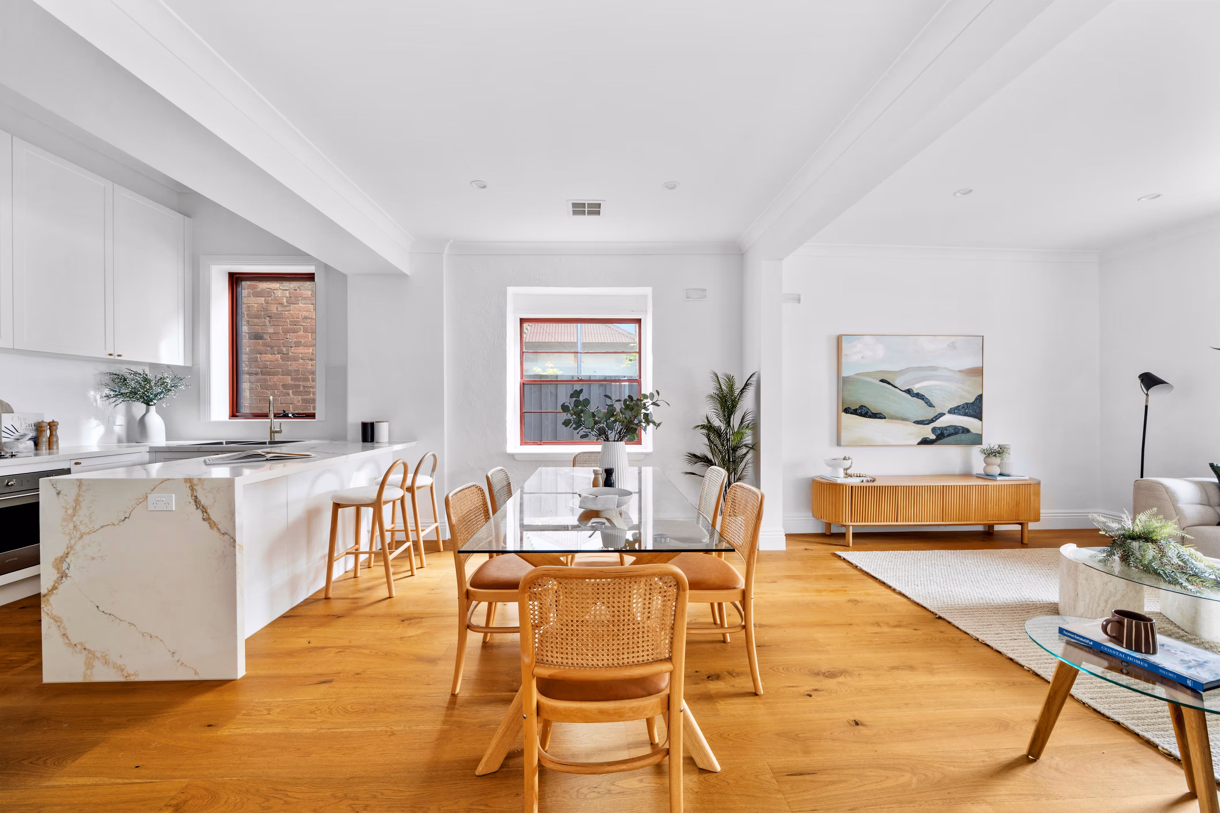 Bright open-plan dining and kitchen area with wood flooring, glass dining table with six wooden chairs, white marble kitchen island, and modern decor.