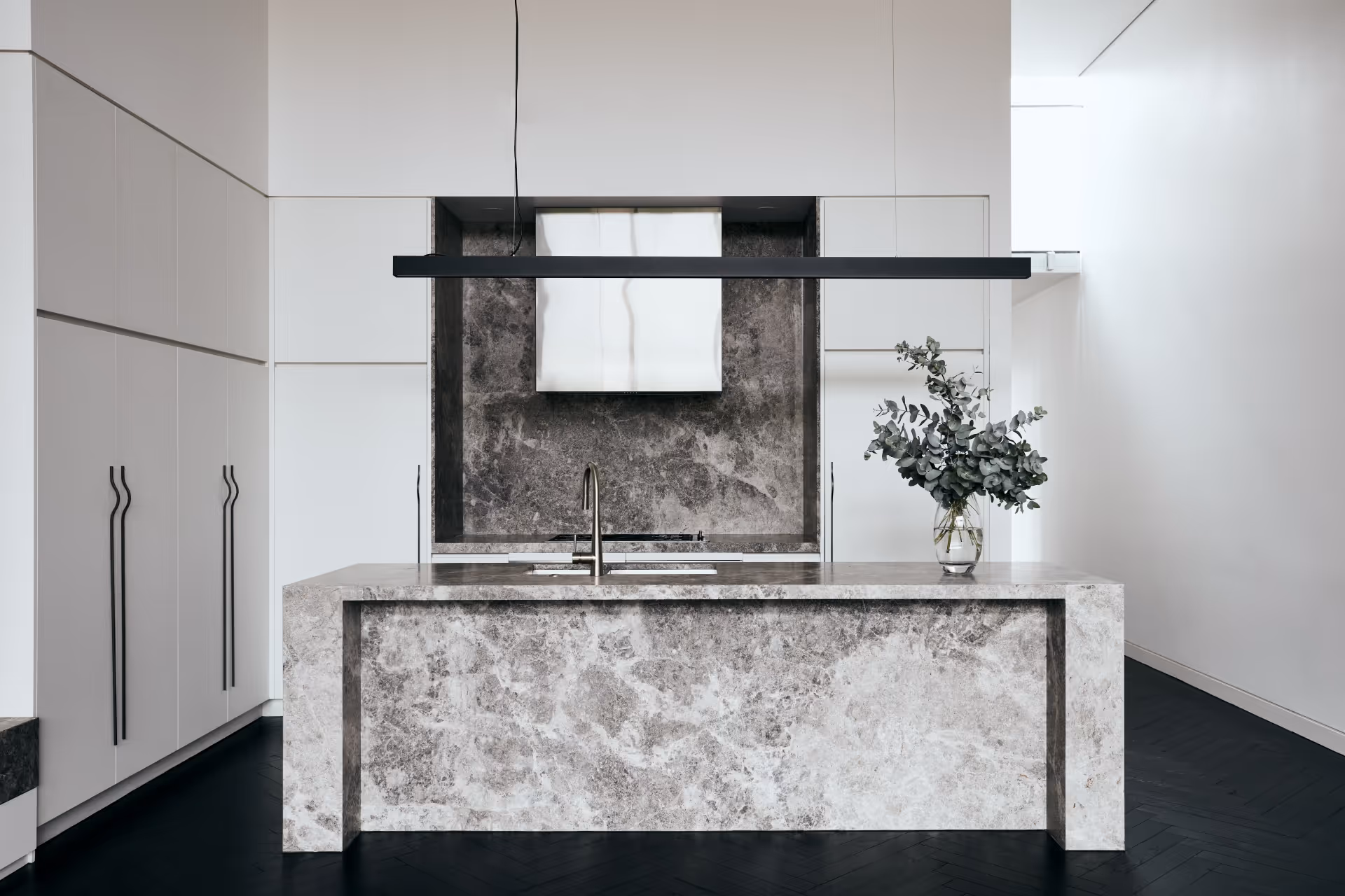 Modern kitchen with a large stone island, integrated sink, sleek white cabinets, and a vase with green foliage.