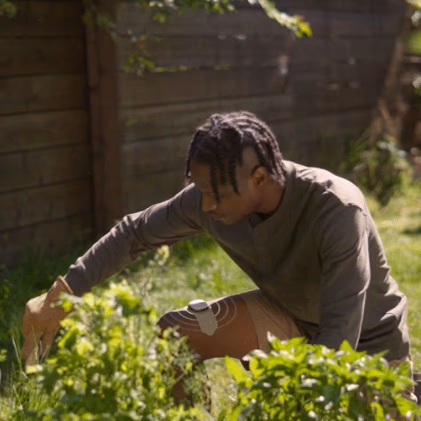 Man with braided hair wearing a gray long-sleeve shirt and beige shorts kneeling near plants in a garden with a small device strapped to his thigh.