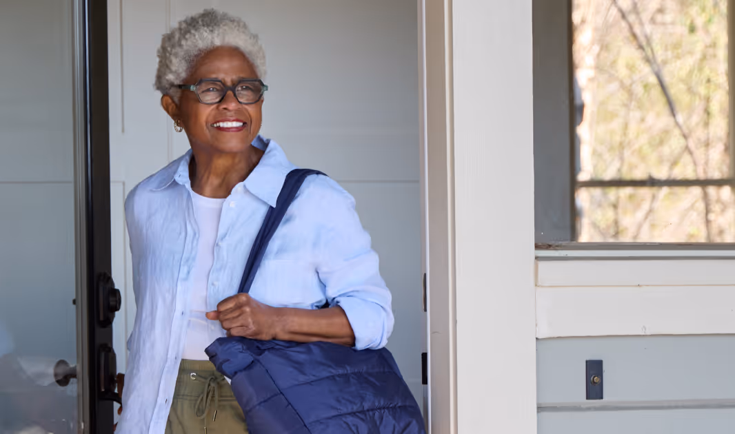 Smiling older woman with gray curly hair wearing glasses, a light blue shirt, and carrying a dark blue bag over her shoulder.