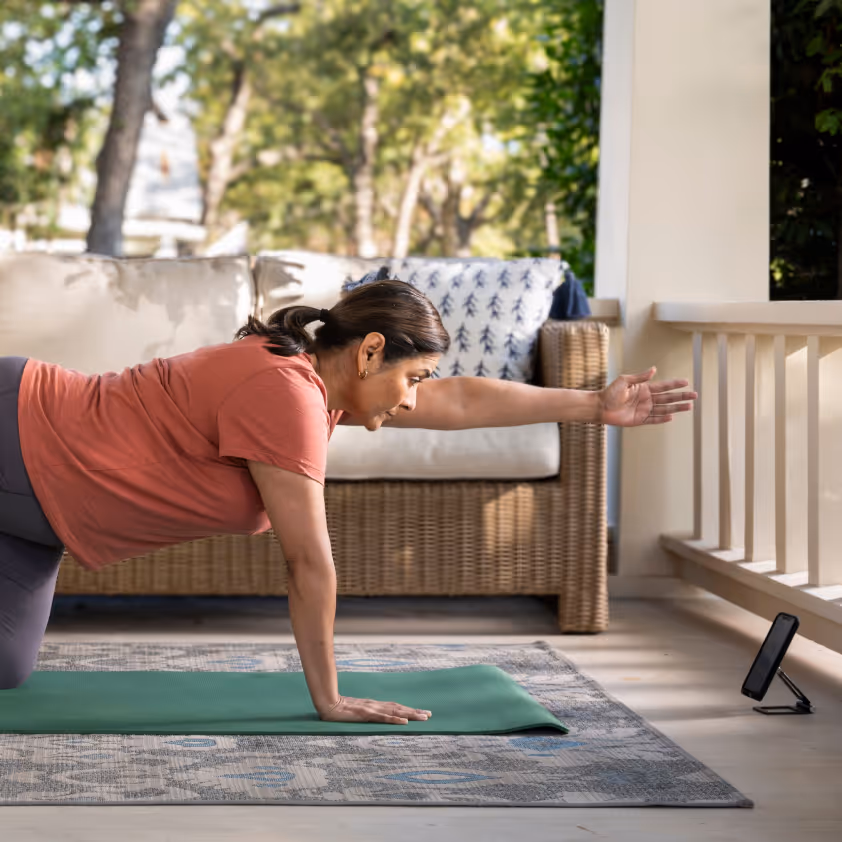 Woman performing a balancing yoga pose on a green mat, following instructions from a smartphone on a porch.