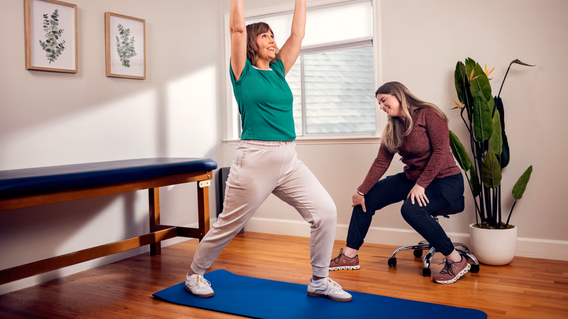 Woman in green shirt doing a yoga stretch on a blue mat while a trainer in a maroon top observes and supports her knee in a bright room.