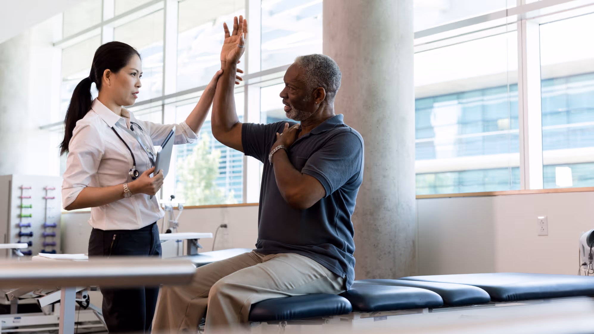 Physical therapist assisting an elderly man seated on a bench with arm exercises in a bright clinic.
